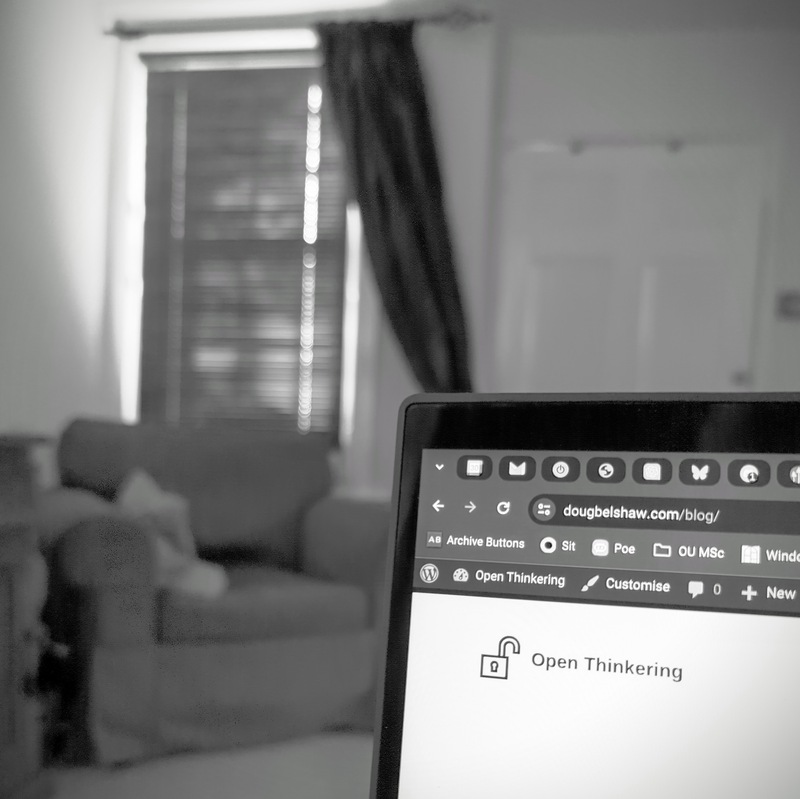 Black and white photograph showing corner of laptop screen in focus with chair, door, and window out of focus in background