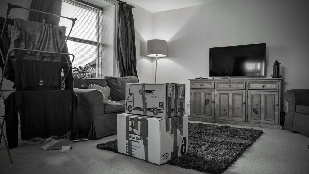 A monochrome photo of a living room with a clothes drying rack, packed moving boxes in the centre, a television on a wooden cabinet, and a cozy armchair, indicating a moving scenario.
