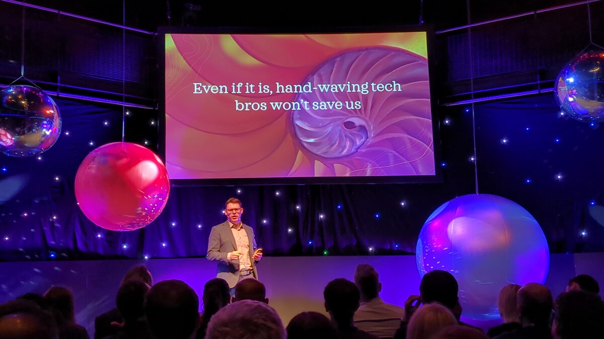 A presenter at a conference with an abstract presentation slide behind him, featuring the text "Even if it is, hand-waving tech bros won't save us", surrounded by large, colorful suspended orbs in a dimly lit room with a starry background.