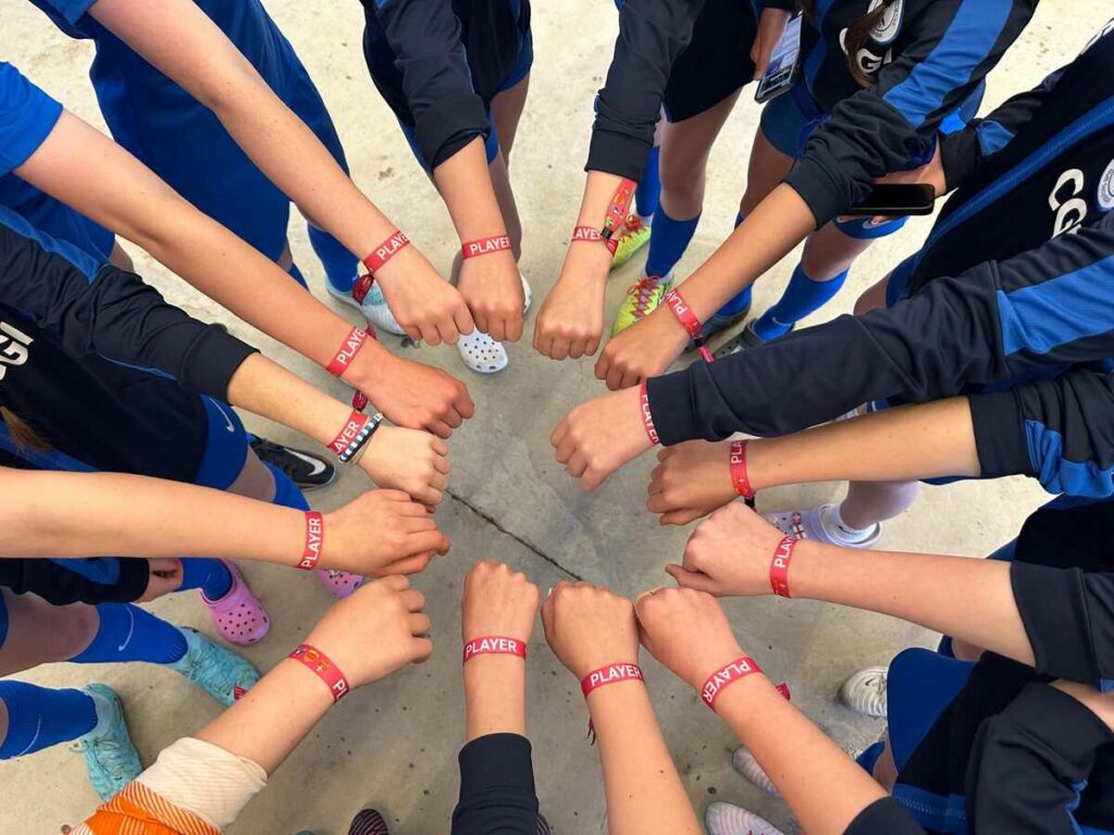 A tight-knit circle of football players' arms reaching in, each with a red 'PLAYER' wristband, symbolizing team unity at a tournament.